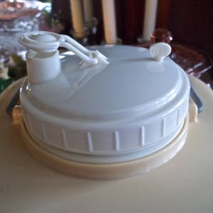 May include: A white plastic lid with a spout and a handle for a large beverage dispenser. The lid is beige on the bottom and has a white, ribbed design.