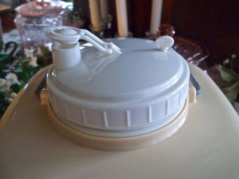 May include: A white plastic lid with a spout and a handle for a large beverage dispenser. The lid is beige on the bottom and has a white, ribbed design.