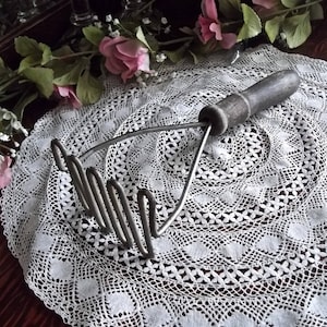 May include: A vintage potato masher with a wooden handle and a coiled metal mashing head. The masher is resting on a white crocheted doily.