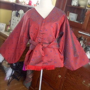 K&ouml;nnte beinhalten: Ein rot-blaues gemustertes Kimono mit langen &Auml;rmeln und einer Sch&auml;rpe. Das Kimono hat einen Fransenbesatz am Saum.