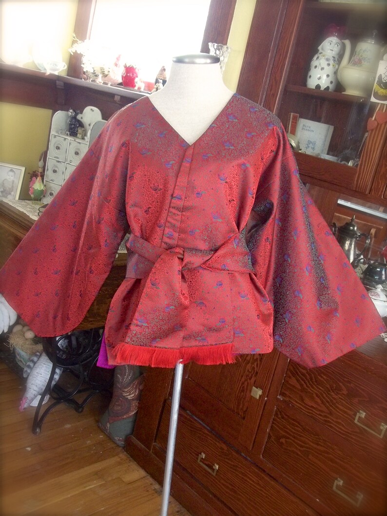 K&ouml;nnte beinhalten: Ein rot-blaues gemustertes Kimono mit langen &Auml;rmeln und einer Sch&auml;rpe. Das Kimono hat einen Fransenbesatz am Saum.