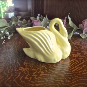 May include: A pale yellow ceramic swan-shaped planter. The swan has a curved neck and a wide, open top for holding plants or flowers. The body features textured, curved lines. The planter is set on a wooden surface.