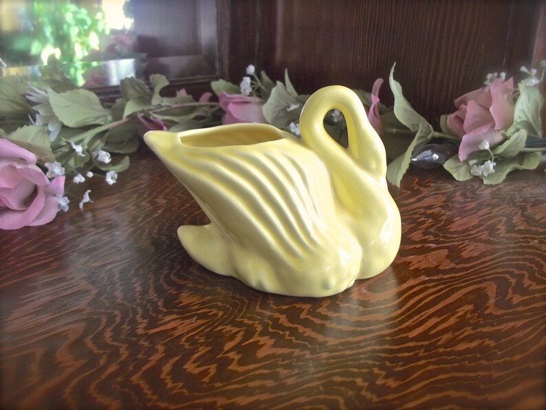 May include: A pale yellow ceramic swan-shaped planter. The swan has a curved neck and a wide, open top for holding plants or flowers. The body features textured, curved lines. The planter is set on a wooden surface.