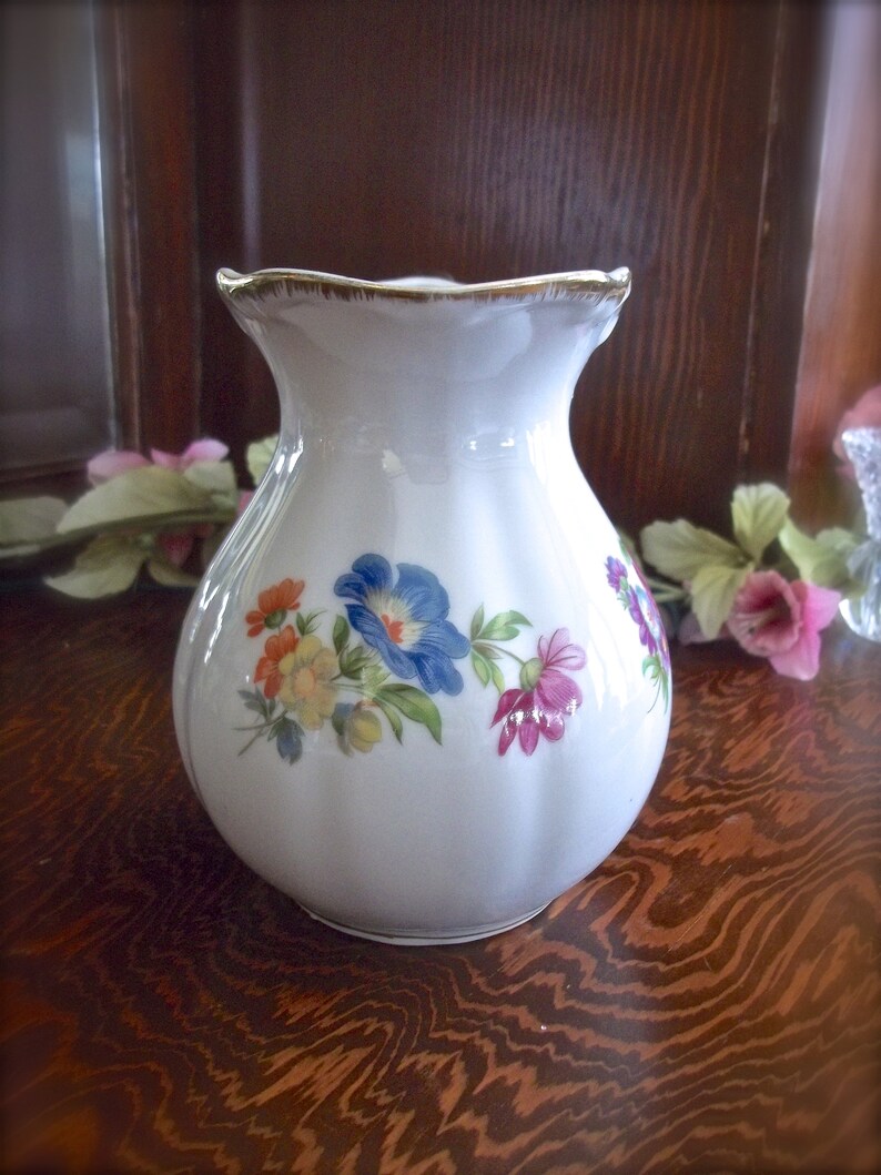 Peut inclure: Un vase en c&eacute;ramique blanche avec un bord festonn&eacute; et un motif floral. Le vase pr&eacute;sente une vari&eacute;t&eacute; de fleurs dans des tons de rose, bleu, jaune et orange.
