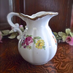 Peut inclure: Un pichet en c&eacute;ramique blanche avec un motif floral. Le pichet a une bordure dor&eacute;e autour du haut et une poign&eacute;e avec un motif floral. Le pichet est pos&eacute; sur une surface en bois.