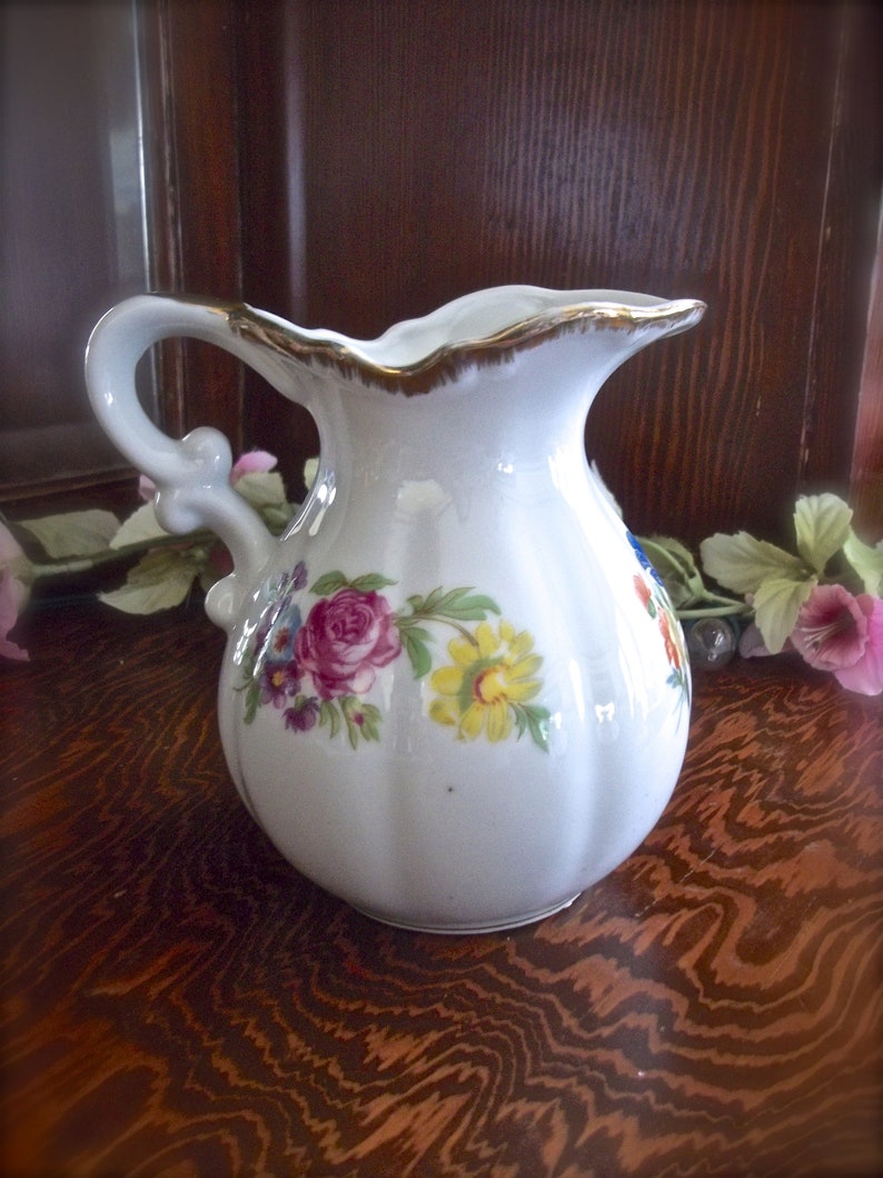 Peut inclure: Un pichet en c&eacute;ramique blanche avec un motif floral. Le pichet a une bordure dor&eacute;e autour du haut et une poign&eacute;e avec un motif floral. Le pichet est pos&eacute; sur une surface en bois.