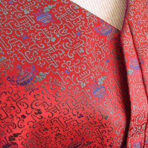K&ouml;nnte beinhalten: Roter Stoff mit einem sich wiederholenden Muster aus blauen, gr&uuml;nen und goldenen stilisierten Zeichen. Der Stoff hat einen violetten Saum.