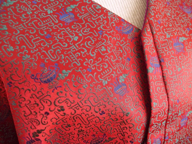 K&ouml;nnte beinhalten: Roter Stoff mit einem sich wiederholenden Muster aus blauen, gr&uuml;nen und goldenen stilisierten Zeichen. Der Stoff hat einen violetten Saum.