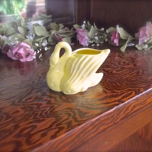 May include: A pale yellow ceramic swan-shaped planter. The swan has a curved neck and open wings, with a small opening at the top for plants. The planter is on a wooden surface with a floral arrangement in the background.