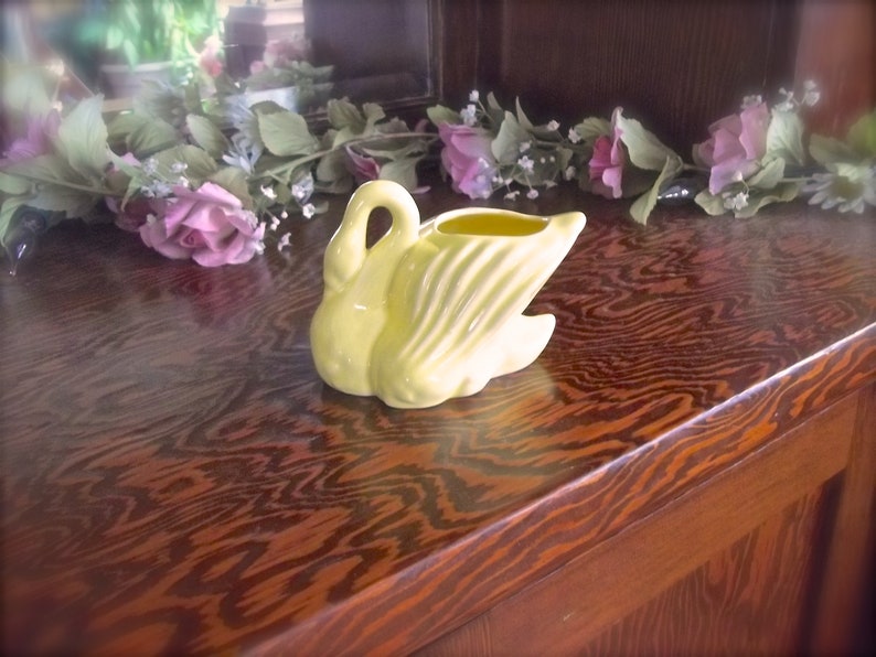May include: A pale yellow ceramic swan-shaped planter. The swan has a curved neck and open wings, with a small opening at the top for plants. The planter is on a wooden surface with a floral arrangement in the background.