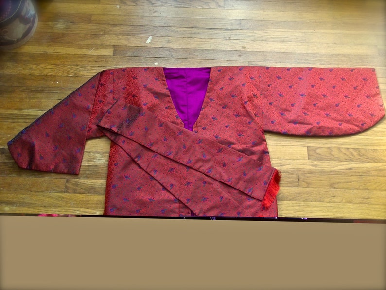 K&ouml;nnte beinhalten: Ein rotes und violettes Kimono mit einem Blumenmuster. Das Kimono hat einen langen, flie&szlig;enden &Auml;rmel und eine Sch&auml;rpe, die sich in der Taille bindet.