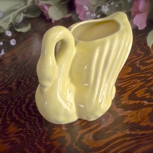 May include: A pale yellow ceramic swan-shaped vase. The swan has a curved neck and a flared tail, with a hollowed-out area for holding flowers or other items. The vase is glossy and sits on a dark wooden surface.