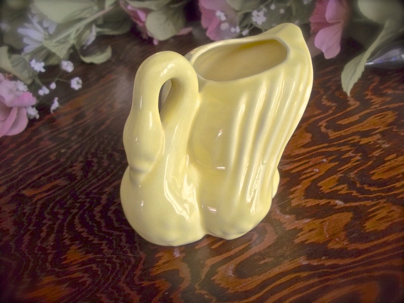 May include: A pale yellow ceramic swan-shaped vase. The swan has a curved neck and a flared tail, with a hollowed-out area for holding flowers or other items. The vase is glossy and sits on a dark wooden surface.