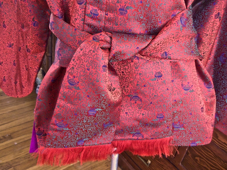 K&ouml;nnte beinhalten: Eine rote Seidenrobe mit einem blauen und gr&uuml;nen Muster. Die Robe hat einen langen Fransen am Saum und einen G&uuml;rtel, der in der Taille gebunden ist.