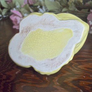 May include: A vintage, yellow ceramic dish with a scalloped edge. The base is a light yellow with a cracked glaze pattern. The rim is white with a slightly aged appearance. The dish is likely a decorative or serving piece.