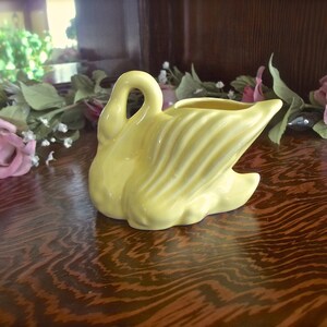 May include: A yellow ceramic swan creamer with a curved neck and detailed wing design. The swan is positioned on a wooden surface, with floral decorations in the background. A decorative piece for the home.