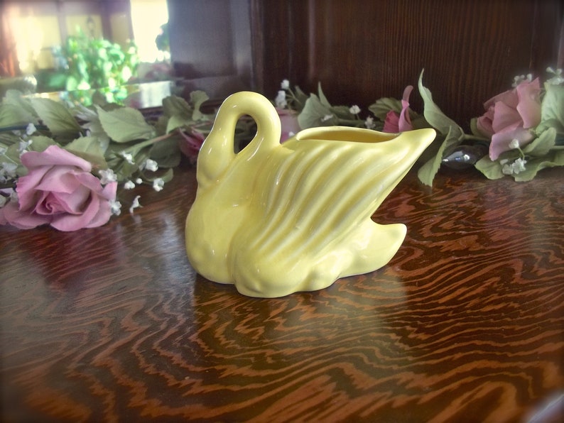 May include: A yellow ceramic swan creamer with a curved neck and detailed wing design. The swan is positioned on a wooden surface, with floral decorations in the background. A decorative piece for the home.