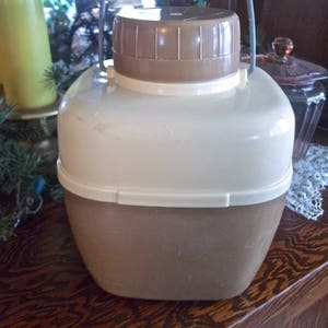 May include: A vintage brown and white plastic insulated beverage pitcher with a lid and a handle. The pitcher has a textured surface and a slightly rounded shape.