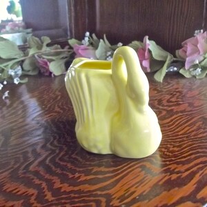 May include: A small, yellow ceramic swan planter with a curved handle. The swan has a textured body and a smooth, rounded base. The planter is sitting on a dark wooden surface.