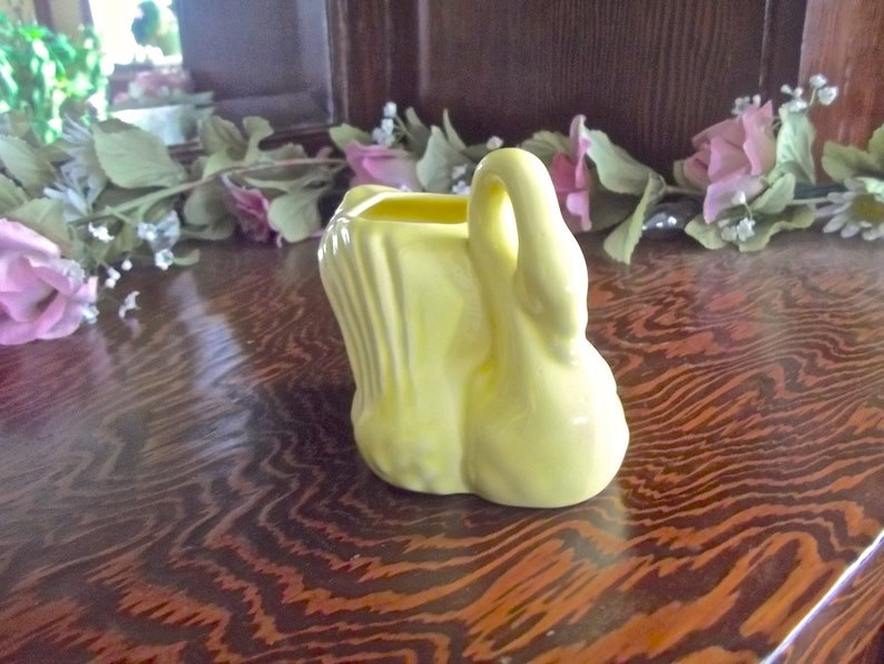 May include: A small, yellow ceramic swan planter with a curved handle. The swan has a textured body and a smooth, rounded base. The planter is sitting on a dark wooden surface.