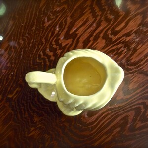 May include: A pale yellow ceramic creamer with a handle, viewed from above. The creamer has a rounded shape with a wide opening and a small spout. The creamer is sitting on a dark brown wooden surface.