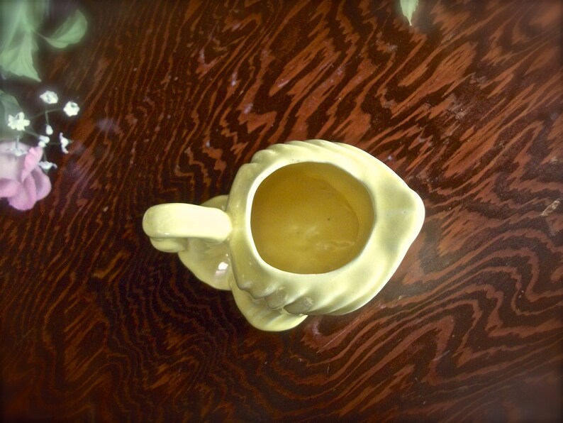 May include: A pale yellow ceramic creamer with a handle, viewed from above. The creamer has a rounded shape with a wide opening and a small spout. The creamer is sitting on a dark brown wooden surface.
