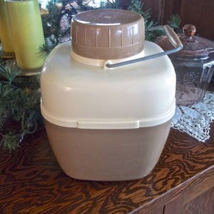 May include: A brown and white insulated beverage container with a lid and a handle. The container has a white top and a brown bottom.