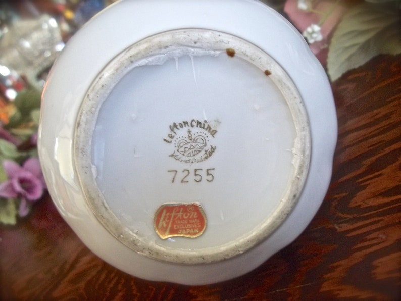 Peut inclure: Vase en porcelaine blanche avec un bord dor&eacute; et un motif floral. Le fond de la vase porte une &eacute;tiquette dor&eacute;e avec le texte "Le Mon China Hand Painted" et le num&eacute;ro "7255". Il y a &eacute;galement une &eacute;tiquette rouge avec le texte "Kiffin Made in Japan Exclusive".