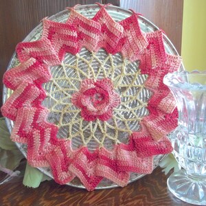 May include: A handmade, circular doily with a central rose design. The doily features a crochet pattern in shades of pink and cream, with a starburst design. The doily is placed on a silver plate.