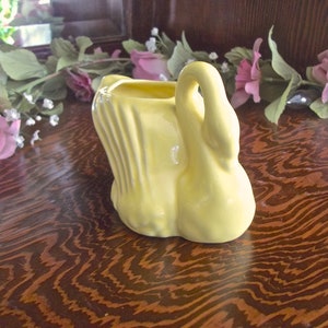 May include: A yellow ceramic swan-shaped planter. The planter has a curved neck and a wide base. The interior is hollow, suitable for holding small plants or flowers. The swan is set on a wooden surface.