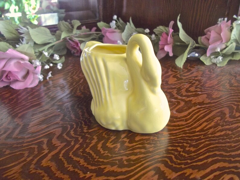 May include: A yellow ceramic swan-shaped planter. The planter has a curved neck and a wide base. The interior is hollow, suitable for holding small plants or flowers. The swan is set on a wooden surface.