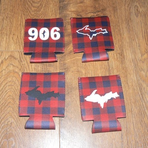 May include: Four red and black buffalo plaid can coolers. Two coolers feature a white outline of the Upper Peninsula of Michigan. One cooler features a black outline of the Upper Peninsula of Michigan. One cooler features the number 906.