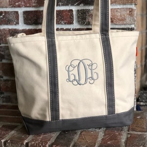 May include: A cream-coloured canvas tote bag featuring dark grey trim along the base and handles. The front of the bag displays a large, embroidered monogram in dark grey thread, consisting of three stylised, interlocking cursive letters.