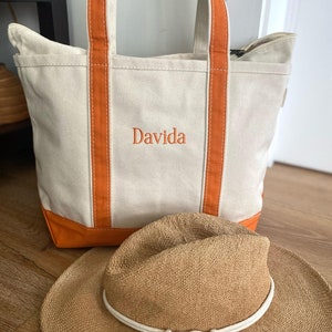 May include: A cream-colored canvas tote bag with orange trim and handles, featuring the embroidered name Davida in orange thread. A tan woven straw hat with a white rope band and knotted tassel detail is positioned in front of the tote bag.