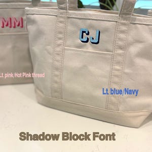 May include: Two canvas tote bags with embroidered initials. One bag features "MM" in hot pink thread, the other "CJ" in light blue and navy thread. The bags have a shadow block font and a front pocket.