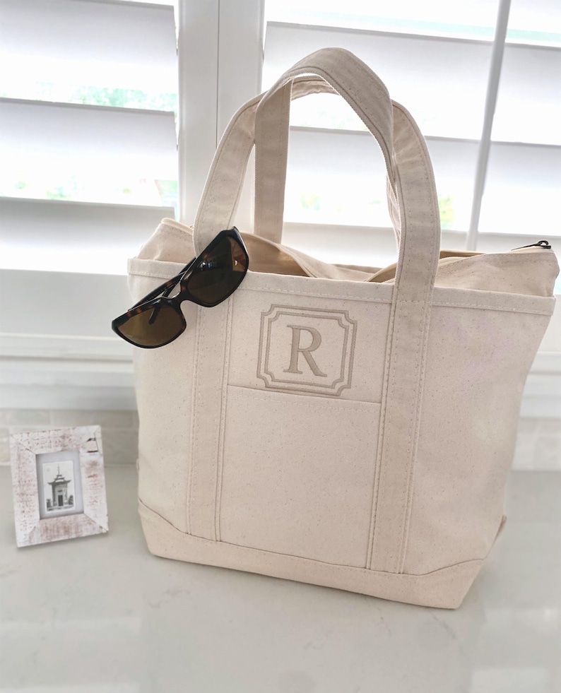 Op de afbeelding: Een cr&egrave;mekleurige canvas draagtas met een centraal geborduurde letter R in een rechthoekig kader. De tas heeft twee stevige handvatten en een ritssluiting aan de bovenkant. Een donkerbruine zonnebril met schildpadpatroon rust op de rand.
