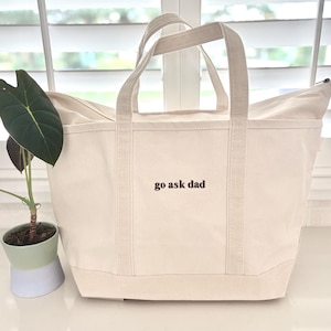May include: A cream-coloured canvas tote bag with two sturdy handles and a zippered top closure. The front of the bag features the text "go ask dad" embroidered in dark brown thread. The bag has a structured base and reinforced vertical stitching.