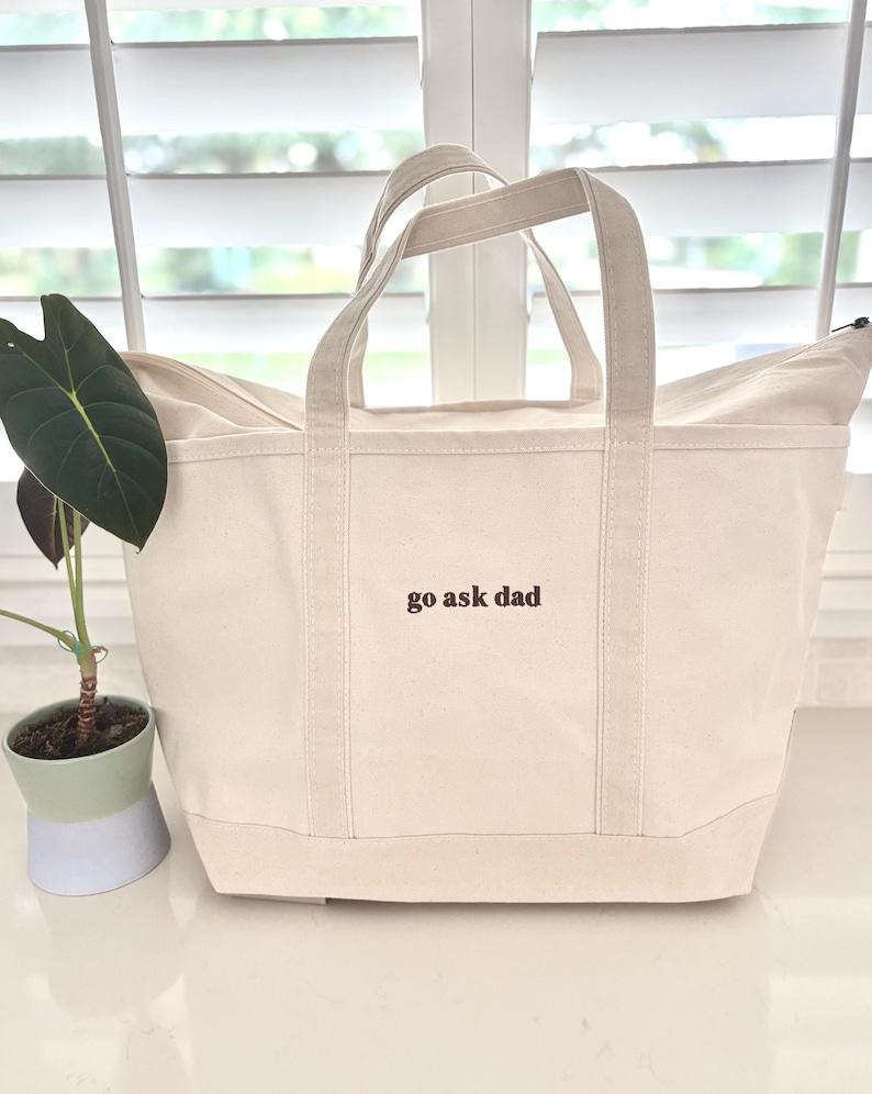 May include: A cream-coloured canvas tote bag with two sturdy handles and a zippered top closure. The front of the bag features the text "go ask dad" embroidered in dark brown thread. The bag has a structured base and reinforced vertical stitching.