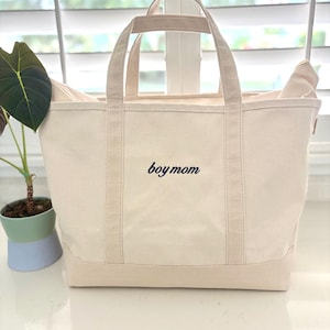 May include: A cream-coloured canvas tote bag with two sturdy handles and a flat base. The front of the bag features the text "boy mom" embroidered in a dark navy blue script font, centred horizontally between the two vertical fabric straps.