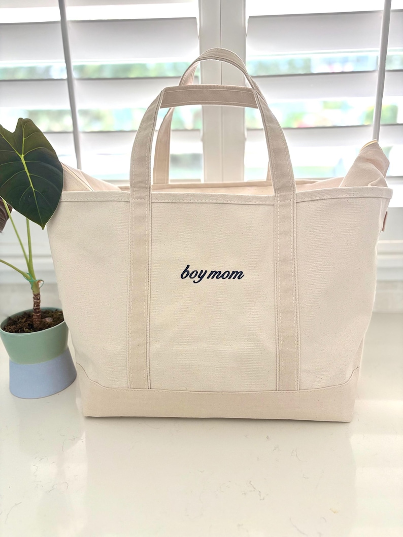 May include: A cream-coloured canvas tote bag with two sturdy handles and a flat base. The front of the bag features the text "boy mom" embroidered in a dark navy blue script font, centred horizontally between the two vertical fabric straps.