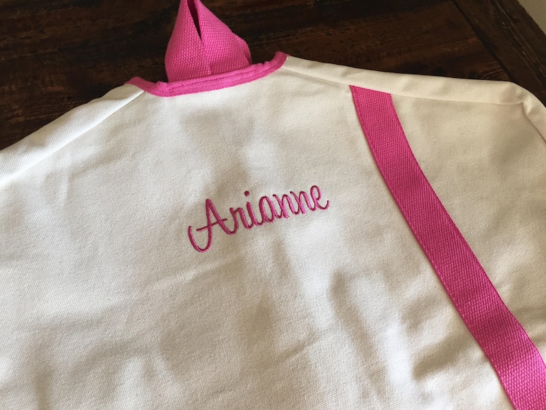 May include: White garment bag with a pink trim and a pink stripe down the side. The bag is personalized with the name "Arianne" embroidered in pink.
