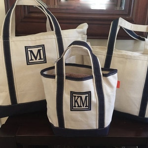 May include: Three canvas tote bags with navy blue trim and handles. The largest bag has the letter "M" embroidered on the front. The medium bag has "KM" embroidered on the front. The smallest bag is plain.