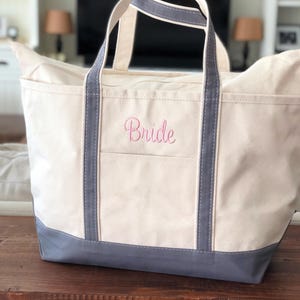 May include: A cream-coloured canvas tote bag with grey trim and handles. The bag features the word Bride embroidered in pink script on the front pocket. The base of the bag is reinforced with a solid grey panel, and the design includes two vertical grey straps extending from the base to the top handles.