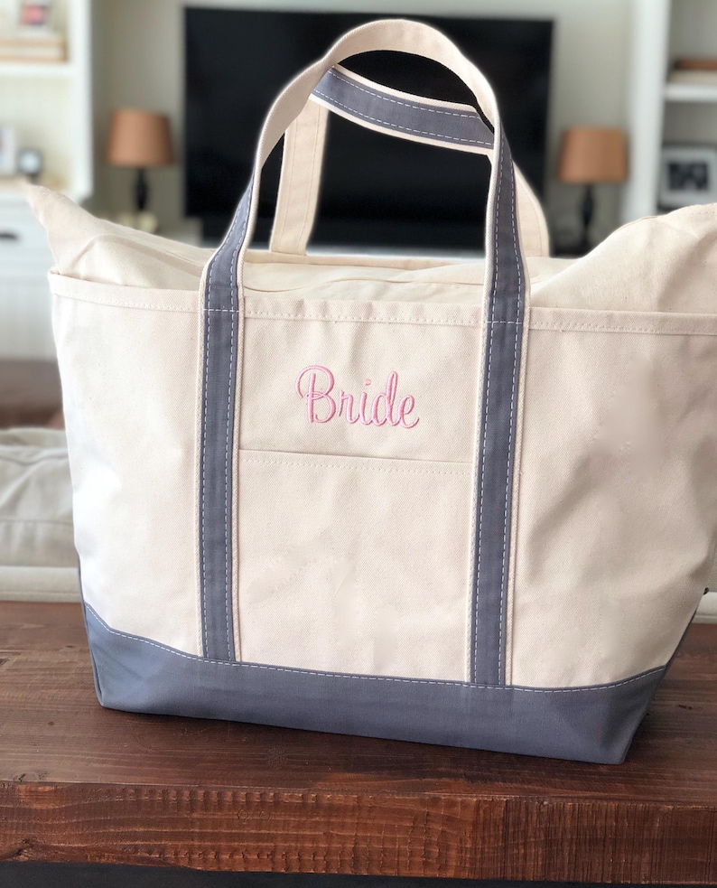 May include: A cream-coloured canvas tote bag with grey trim and handles. The bag features the word Bride embroidered in pink script on the front pocket. The base of the bag is reinforced with a solid grey panel, and the design includes two vertical grey straps extending from the base to the top handles.
