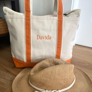 May include: A cream-coloured canvas tote bag with orange trim and handles, featuring the name Davida embroidered in orange thread on the front. A tan woven straw hat with a white rope band and tassel detail is positioned in front of the bag.