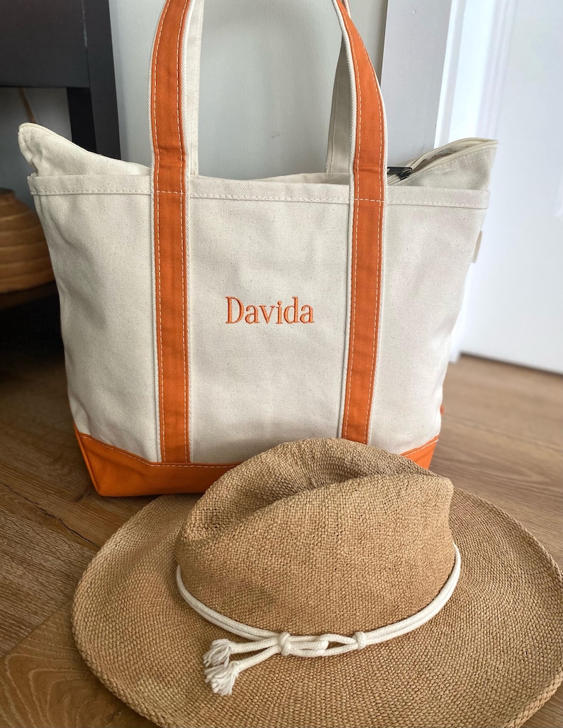 May include: A cream-coloured canvas tote bag with orange trim and handles, featuring the name Davida embroidered in orange thread on the front. A tan woven straw hat with a white rope band and tassel detail is positioned in front of the bag.