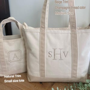 May include: Two off-white canvas tote bags with beige trim. The larger bag features the embroidered monogram SHv in champagne thread and text reading Beige Trim, Champagne thread color, Medium Size. The smaller bag has an embroidered A and text reading Natural Trim, Small size tote.