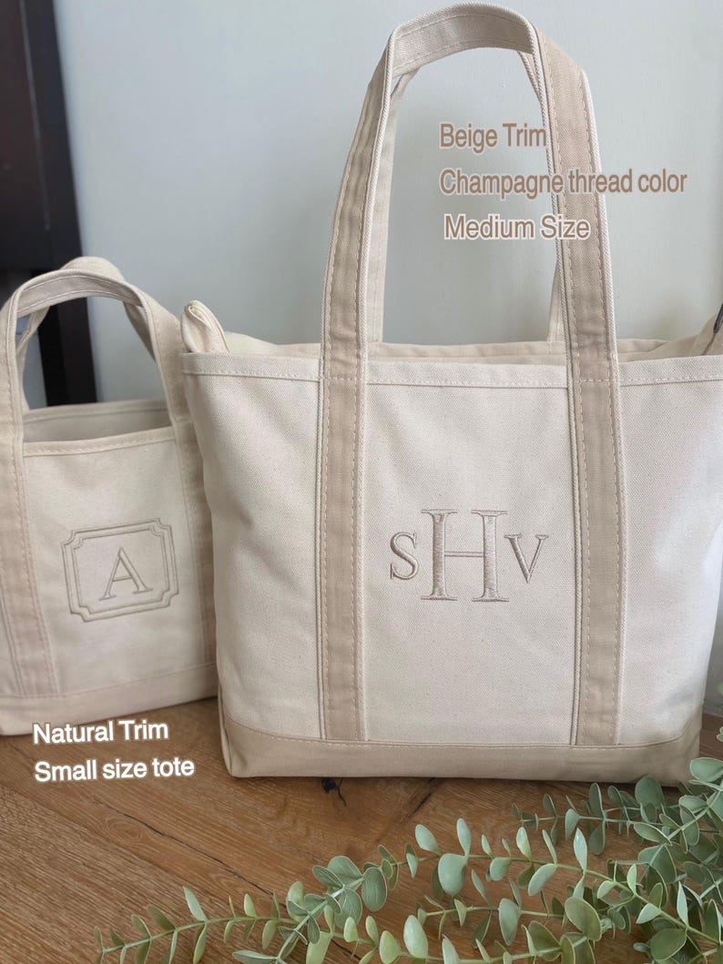 May include: Two off-white canvas tote bags with beige trim. The larger bag features the embroidered monogram SHv in champagne thread and text reading Beige Trim, Champagne thread color, Medium Size. The smaller bag has an embroidered A and text reading Natural Trim, Small size tote.