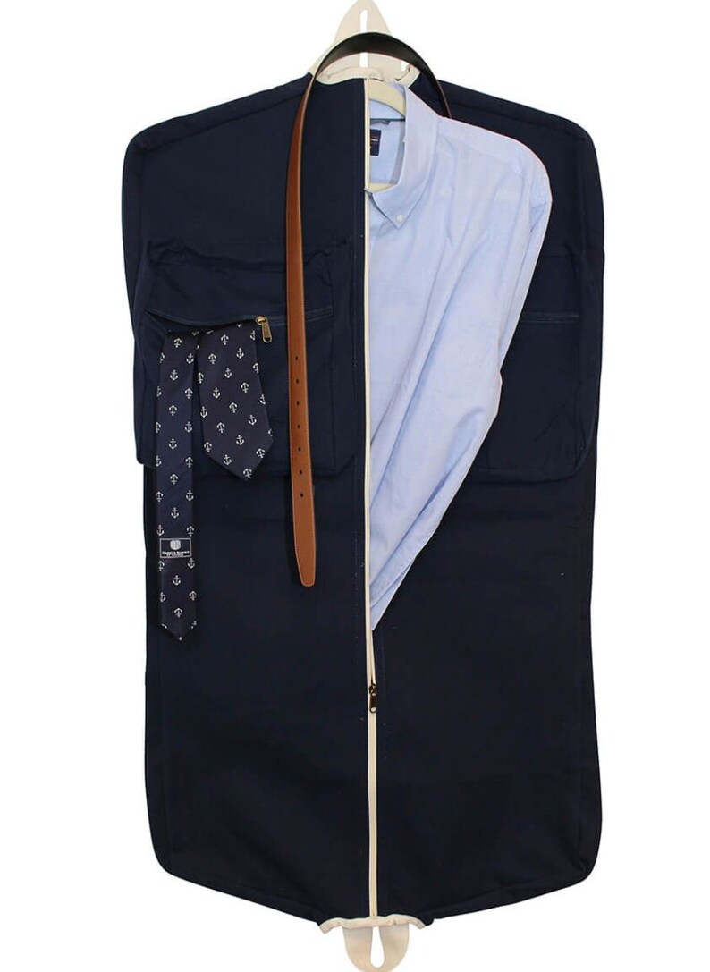 May include: A navy blue garment bag with a white zipper and a brown leather belt. The bag has a pocket on the front with a tie and a pocket on the back with a shirt.