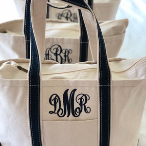 Op de afbeelding: Een natuurlijke canvas tas met een zwarte onderkant, zwarte handvatten en een zwarte ritssluiting. De voorkant van de tas is voorzien van een zwart geborduurd monogram met de letters DMR in een decoratief scriptlettertype boven een zak.